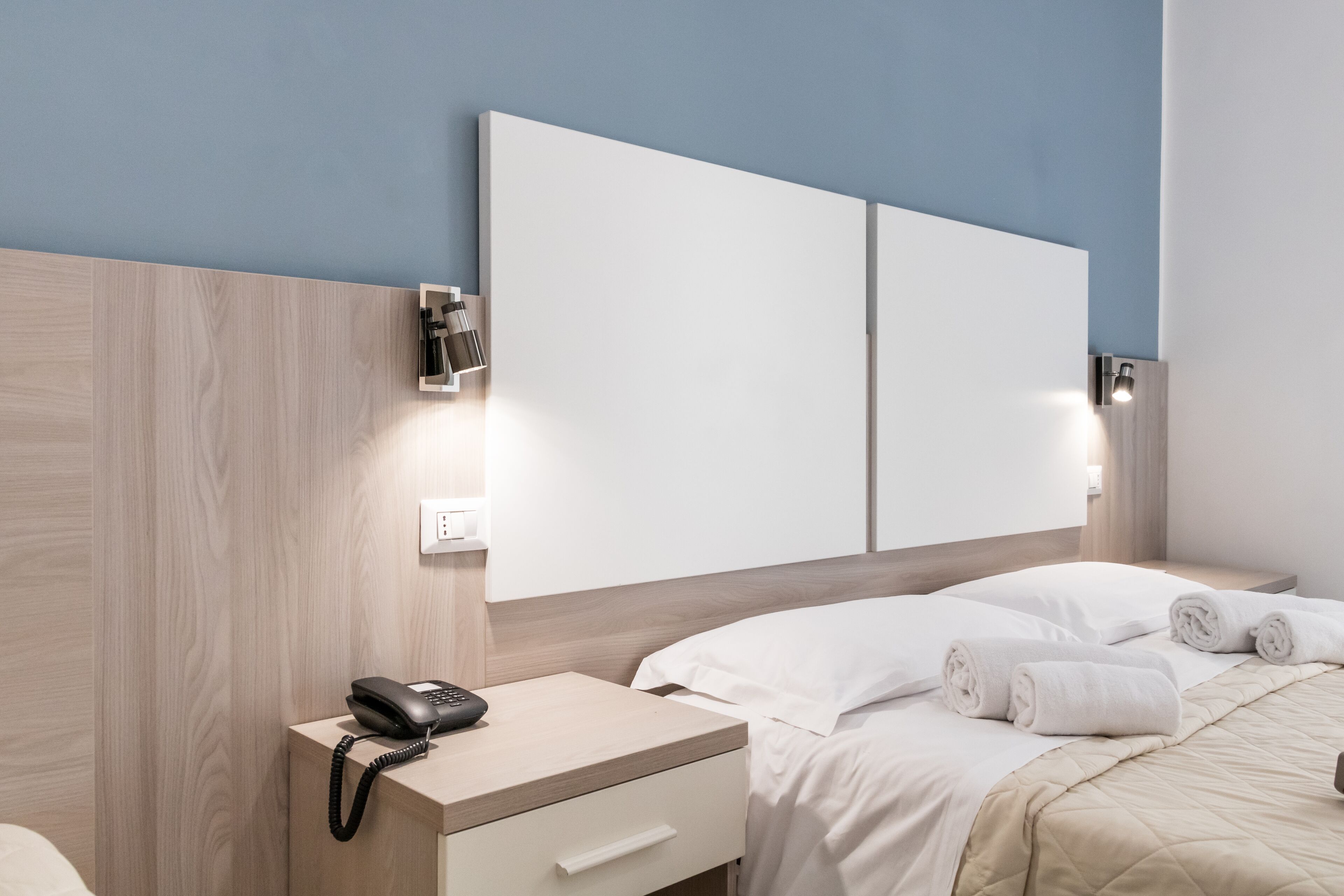 Ett modernt hotellrum med en sänggavel i trä, en telefon vid sängen, en stor säng med vita sängkläder och en väggmonterad lampa.