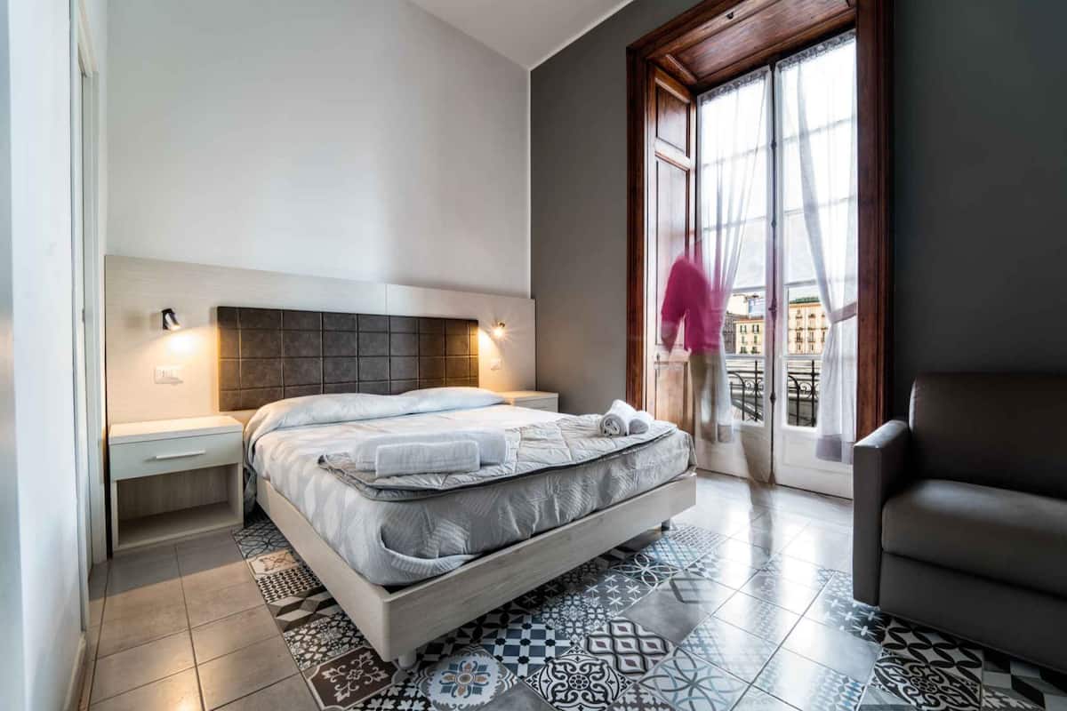 A modern bedroom with a large bed, bedside tables, a patterned rug, and a view of buildings through a window.