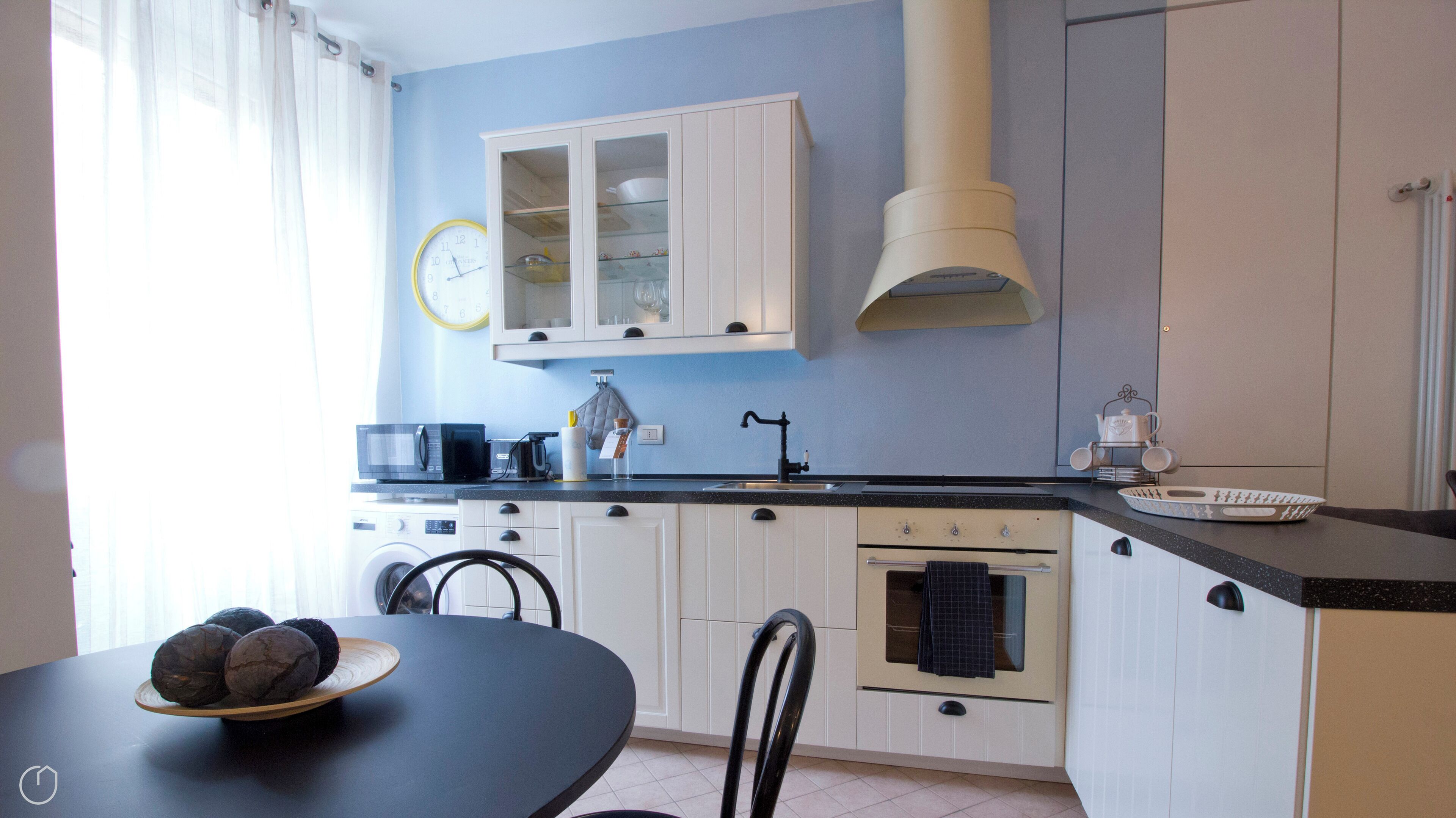Une cuisine avec des meubles blancs, un plan de travail noir, un four à micro-ondes, un évier et une horloge au mur.