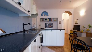 Une cuisine moderne avec un plan de travail noir, des armoires blanches et un four encastré. On y trouve un coin repas avec une table et des chaises, ainsi qu’une bibliothèque contenant des livres et des objets décoratifs.