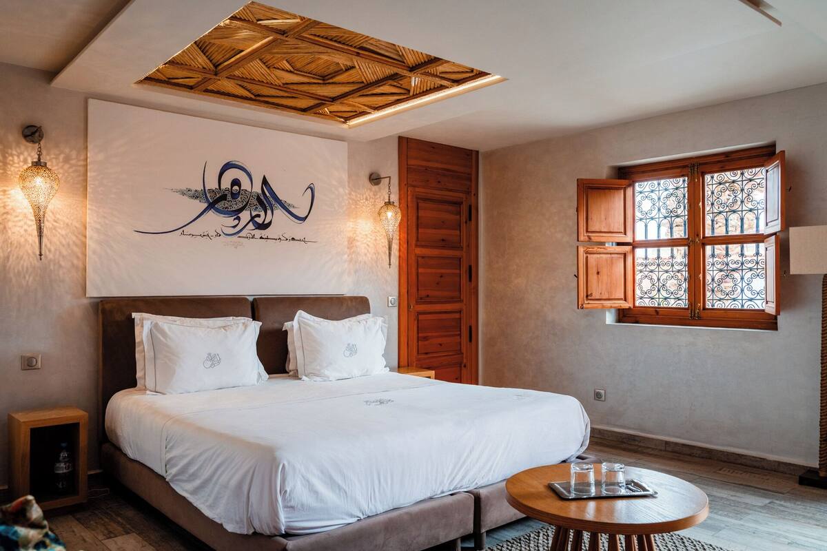 A bedroom with a large bed, a wooden door, a window with decorative iron bars, and a ceiling with a geometric pattern.
