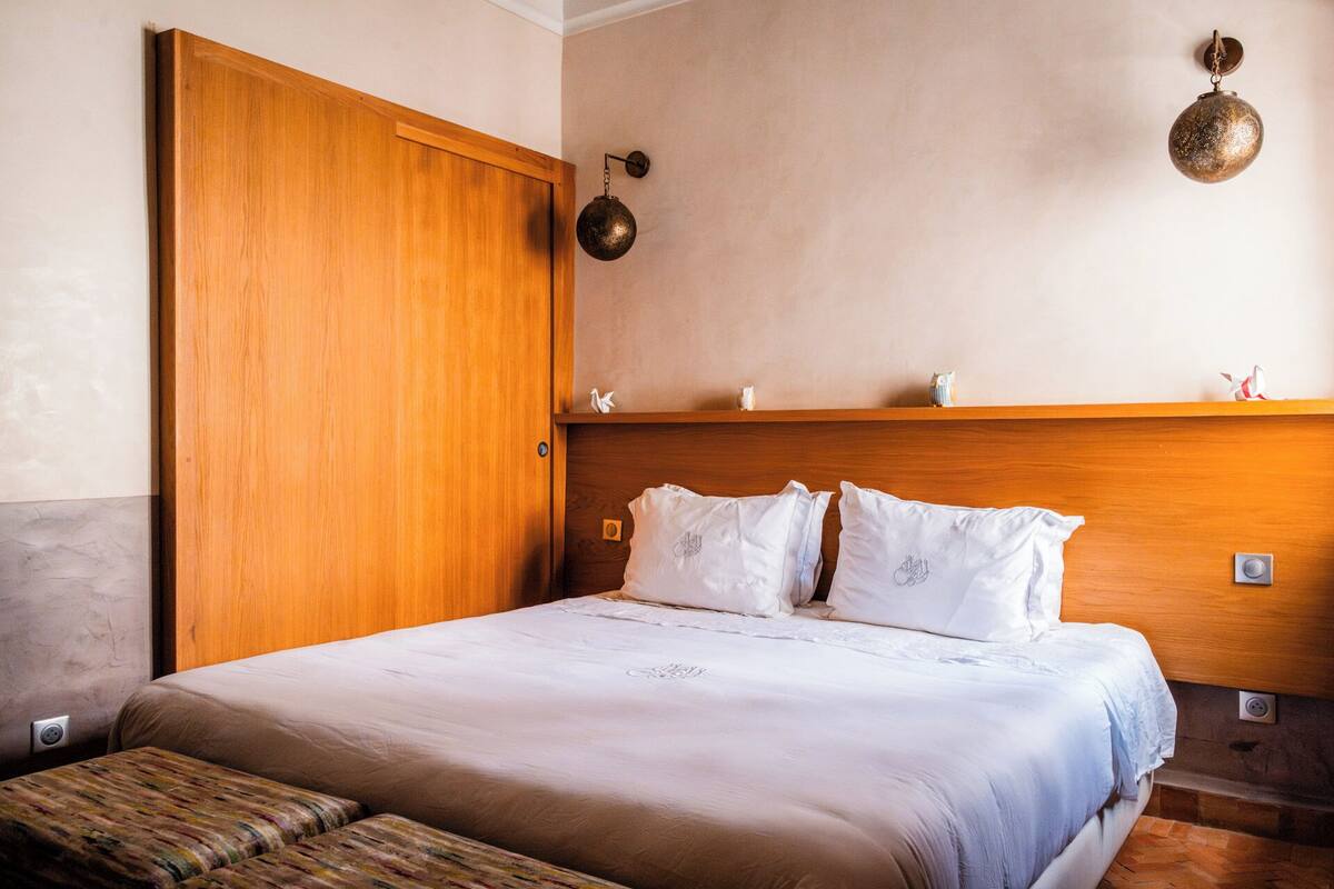 A bedroom with a wooden door, a bed with white bedding, a headboard, and a shelf with decorative items.