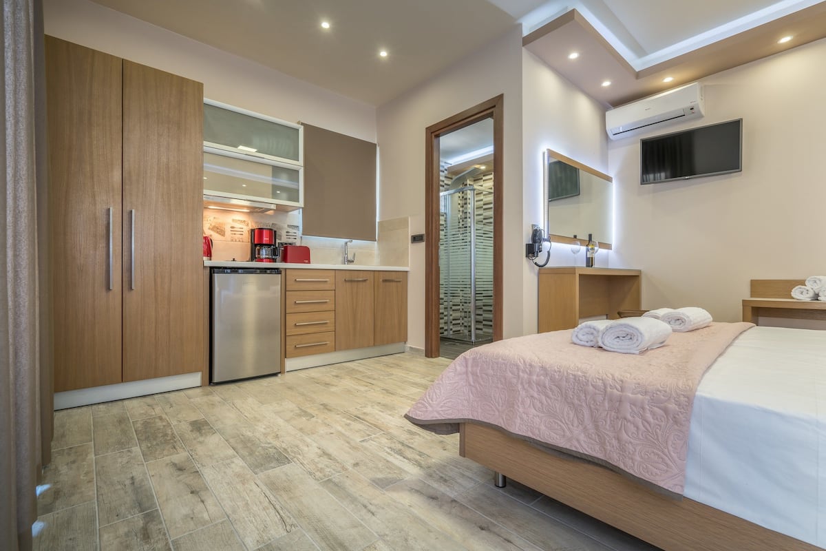 A modern hotel room with a kitchenette, a bed with a pink bedspread, a TV mounted on the wall, and an air conditioning unit.