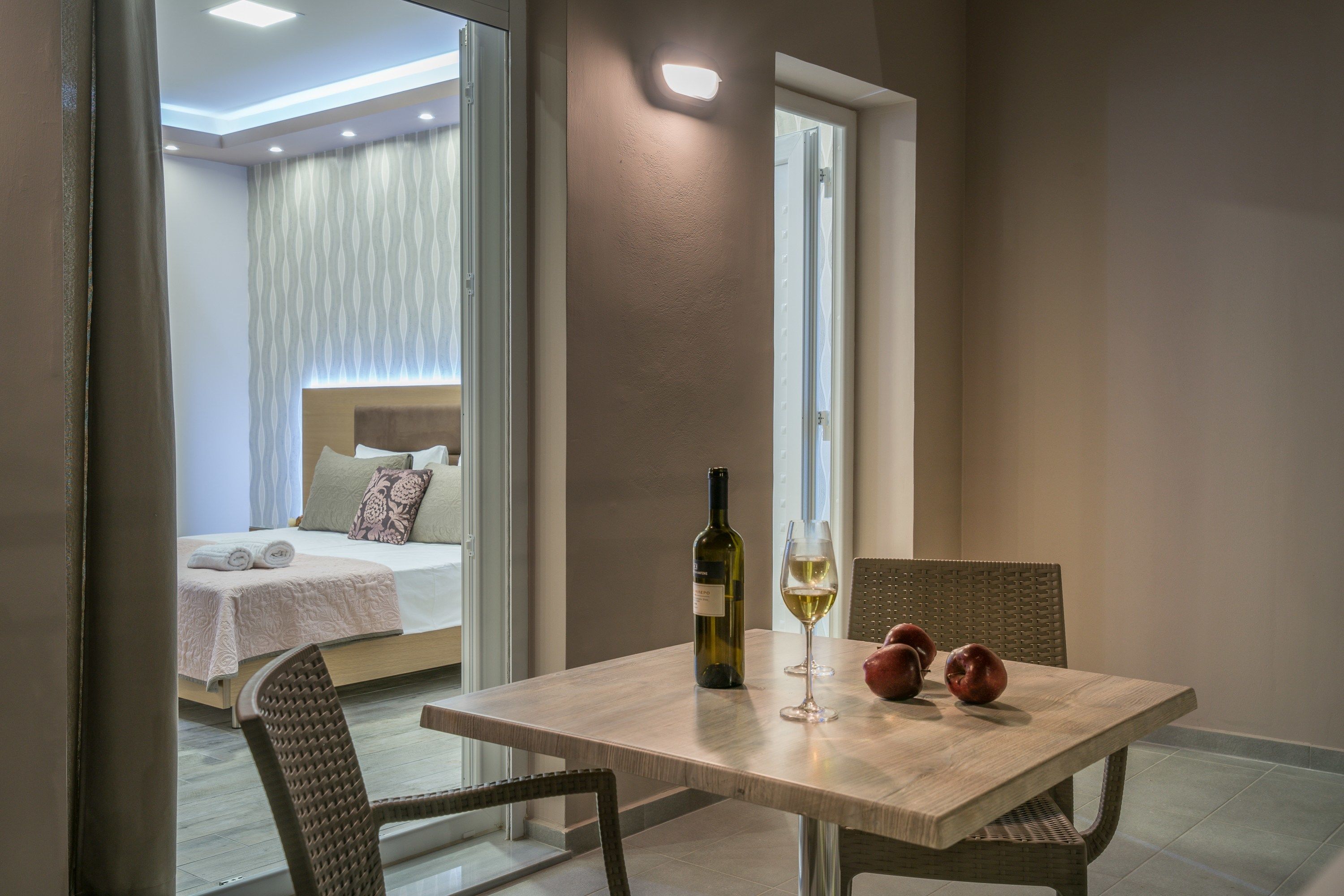 A modern bedroom with a bed, a bedside table with a wine bottle and glass, and a dining area with chairs and a table.