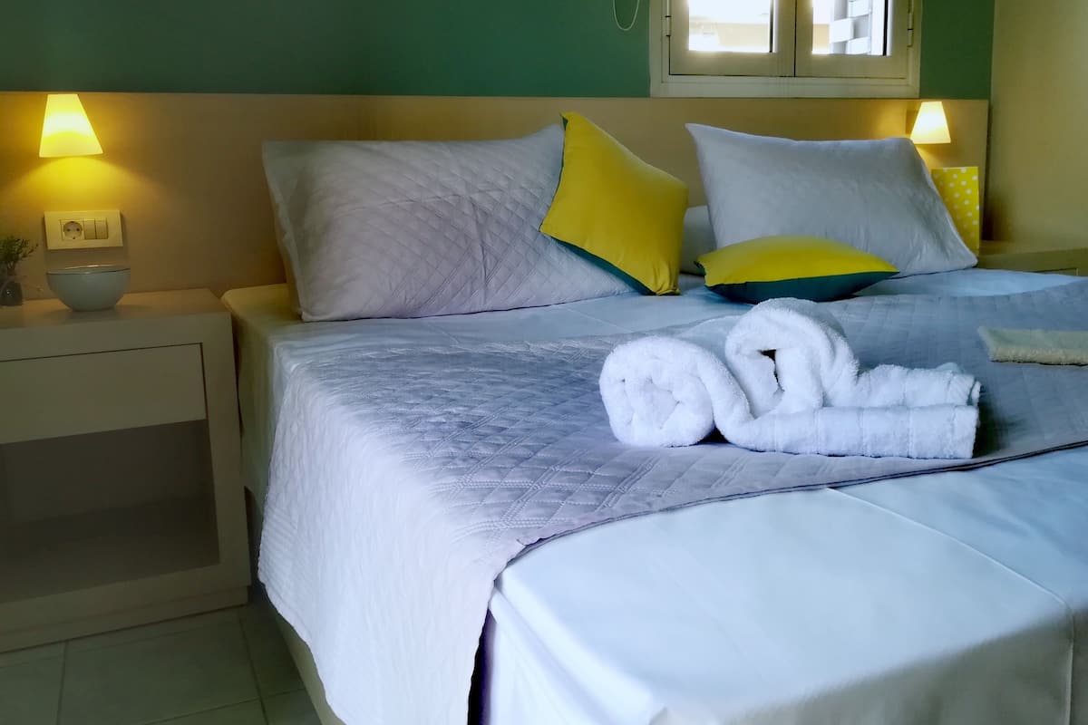A neatly made bed with white and yellow pillows, a bowl on the nightstand, and a window with curtains.
