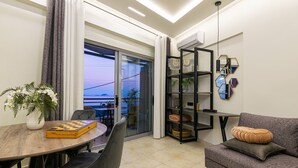 A modern living room with a wooden dining table, black chairs, a grey sofa, and a glass door leading to a balcony. - Mouria Studios & Apartments (Zakynthos)