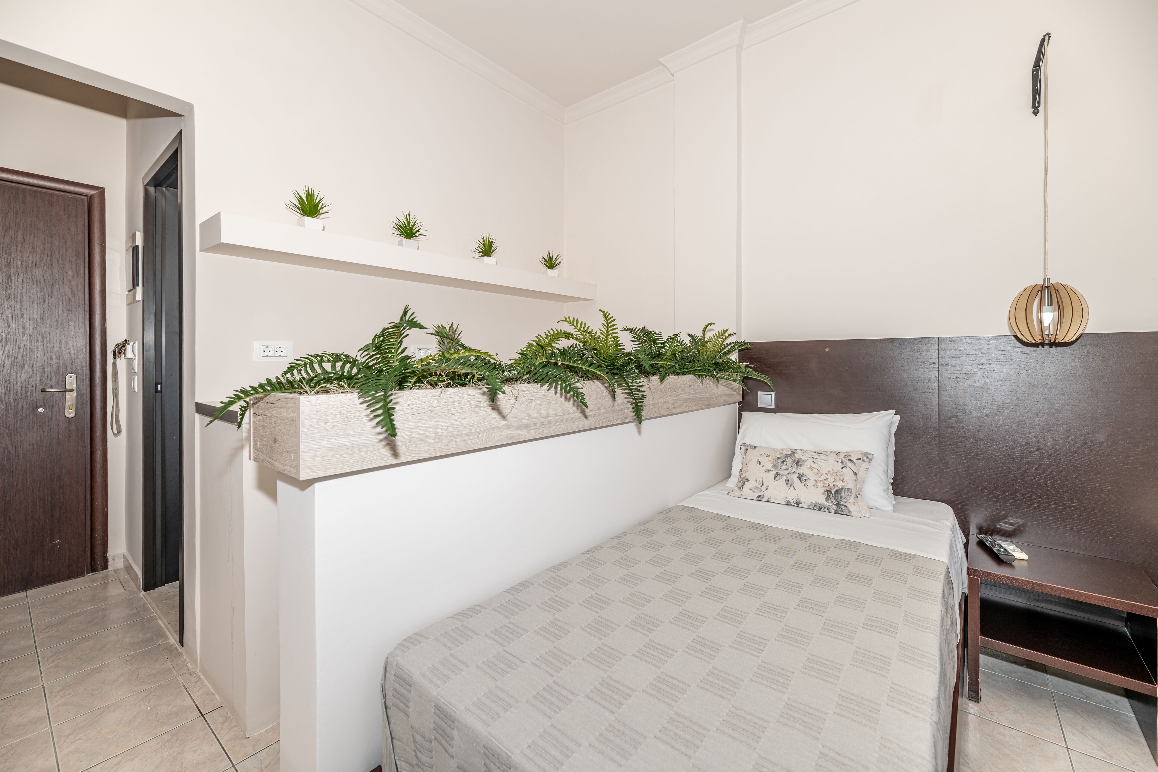 A bedroom with a bed, a nightstand, a wall-mounted shelf with plants, and a hanging light fixture.