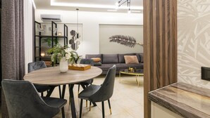 A modern living room with a round wooden dining table, dark upholstered chairs, a grey sofa, a coffee table, and decorative items including a large feather art piece on the wall. - Mouria Studios & Apartments (Zakynthos)