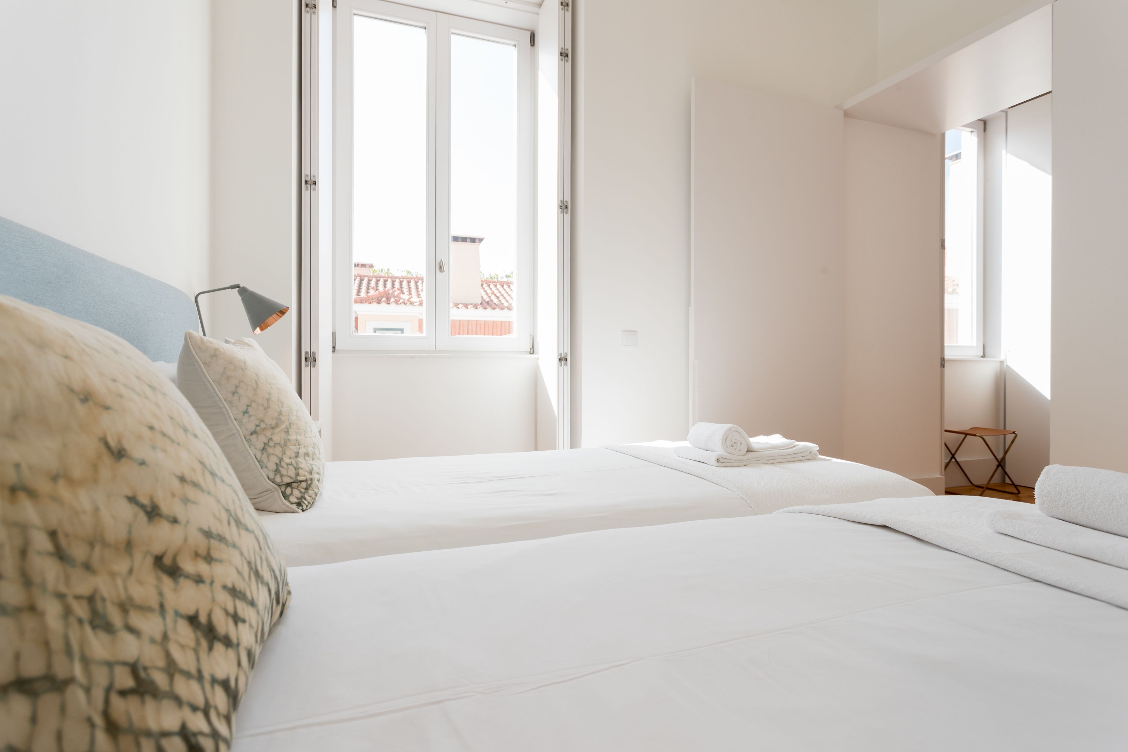 A hotel room with two beds, a window with a view of buildings, a small table, and a chair.