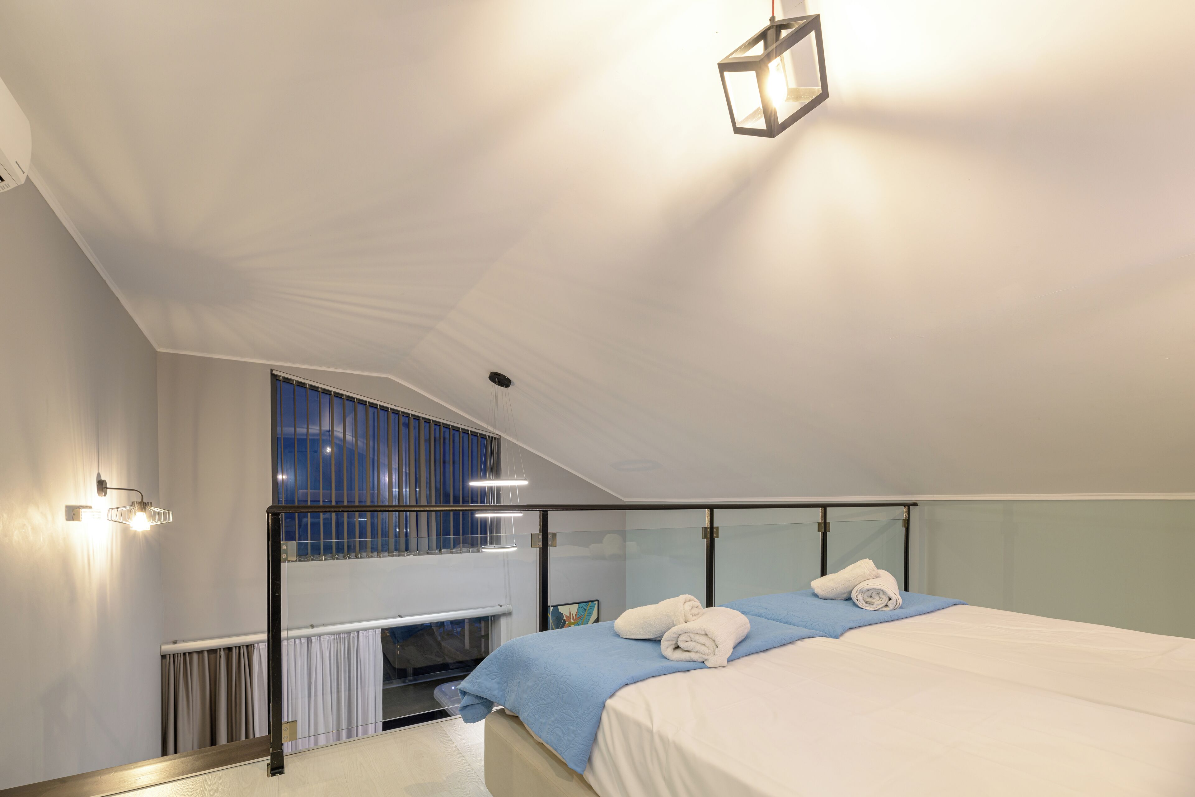 A modern hotel room with a large bed, a glass partition, a view of the city, and a balcony with a railing.