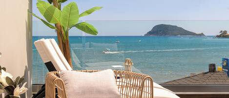 A balcony with a wicker chair and a table, overlooking a calm sea with boats and a distant island.