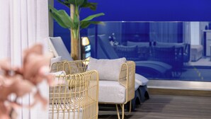Uma sala de estar moderna com um sofá, poltronas, uma planta grande em vaso e vista para uma piscina através de um muro de vidro.