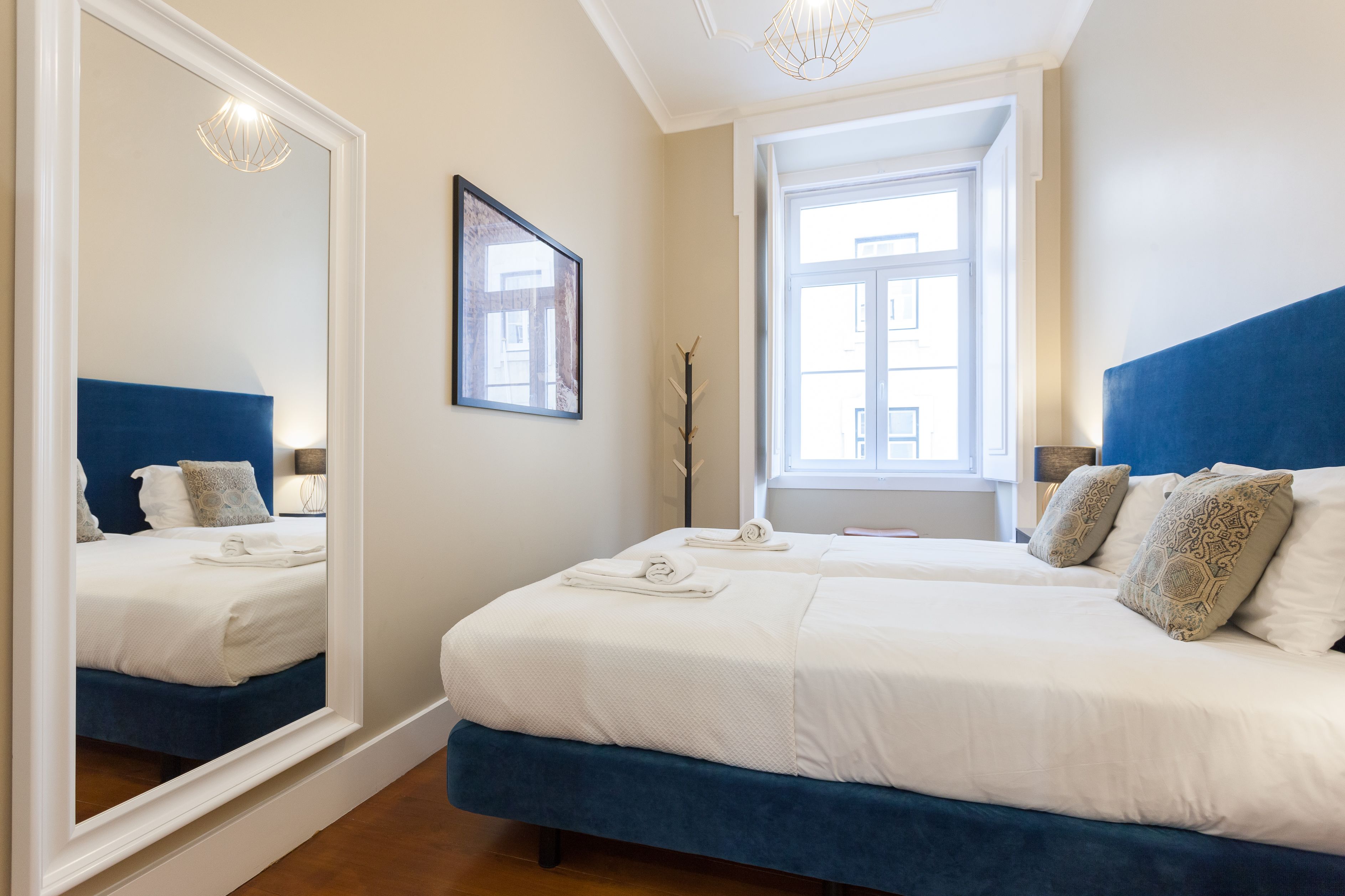 A hotel room with a large mirror, a bed with white bedding, a blue headboard, and a window with natural light.