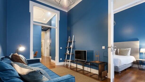 A modern living room with a blue sofa, a TV on a stand, and a bed with a white headboard.