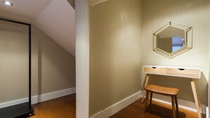 A room with a wooden floor, a white desk with drawers, a small wooden stool, and a hexagonal mirror on the wall.