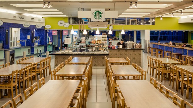 A spacious dining area with multiple wooden tables and chairs, a central counter with a coffee machine, and a bright, modern interior design. - DCU Rooms - Campus Accommodation (Dublin)