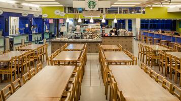 A spacious dining area with multiple wooden tables and chairs, a central counter with a coffee machine, and a bright, modern interior design.