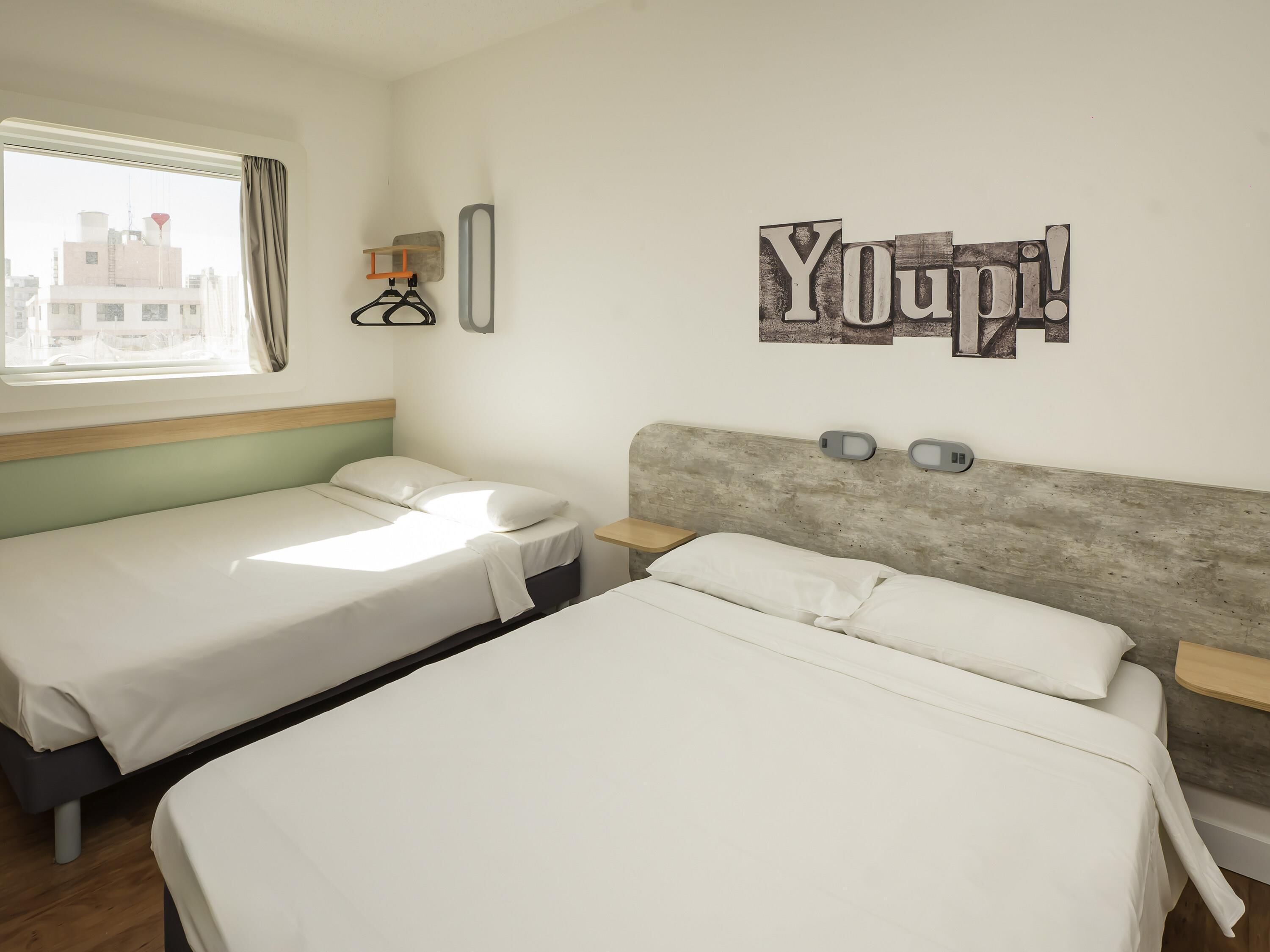 Quarto de hotel com duas camas, uma janela com cortinas, prateleira na parede com um relógio e um aviso com a inscrição 'Youpi!'.