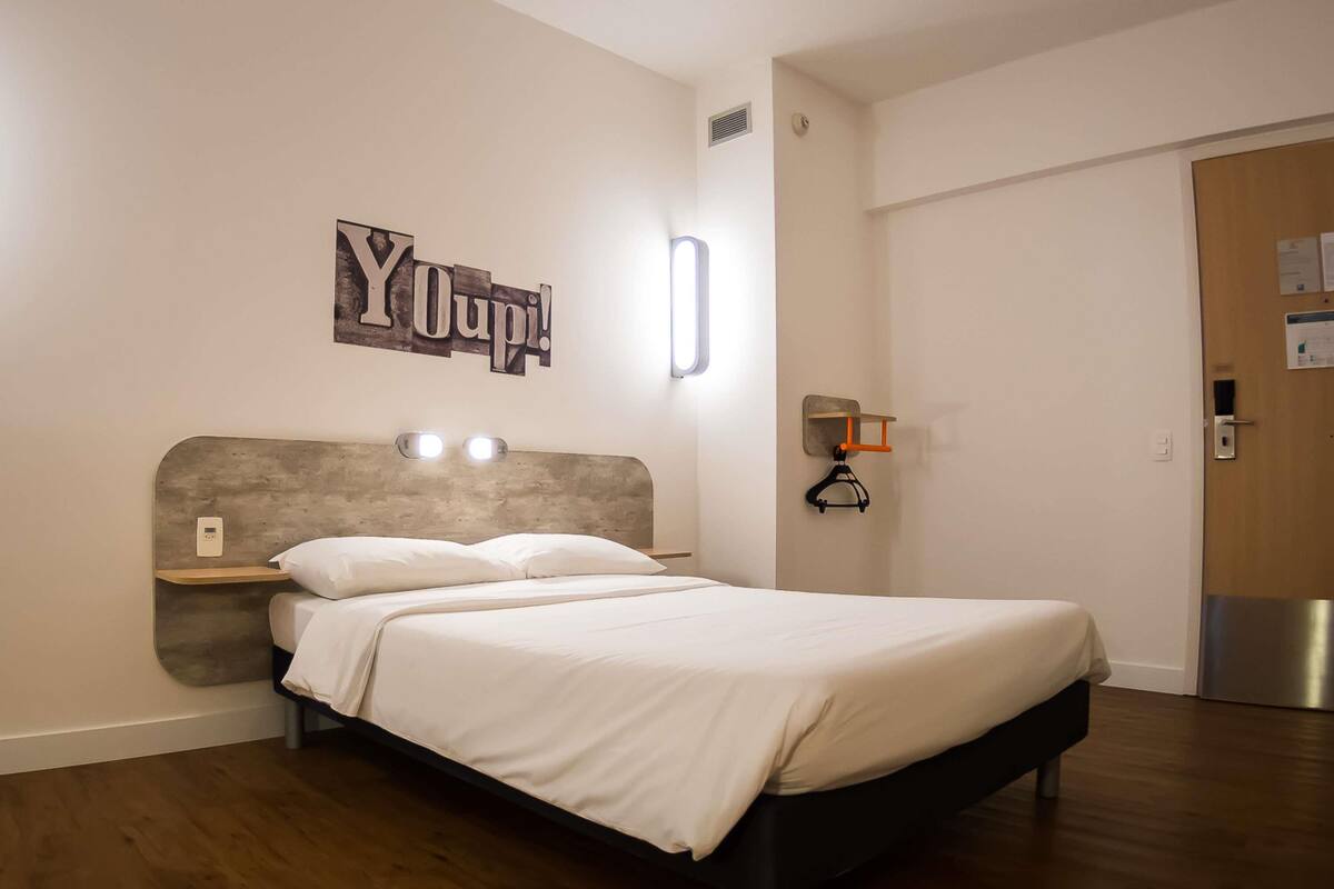 Une chambre d’hôtel moderne, équipée d’un lit, d’une tête de lit avec des lampes, d’une étagère fixée au mur et d’une porte avec une poignée.