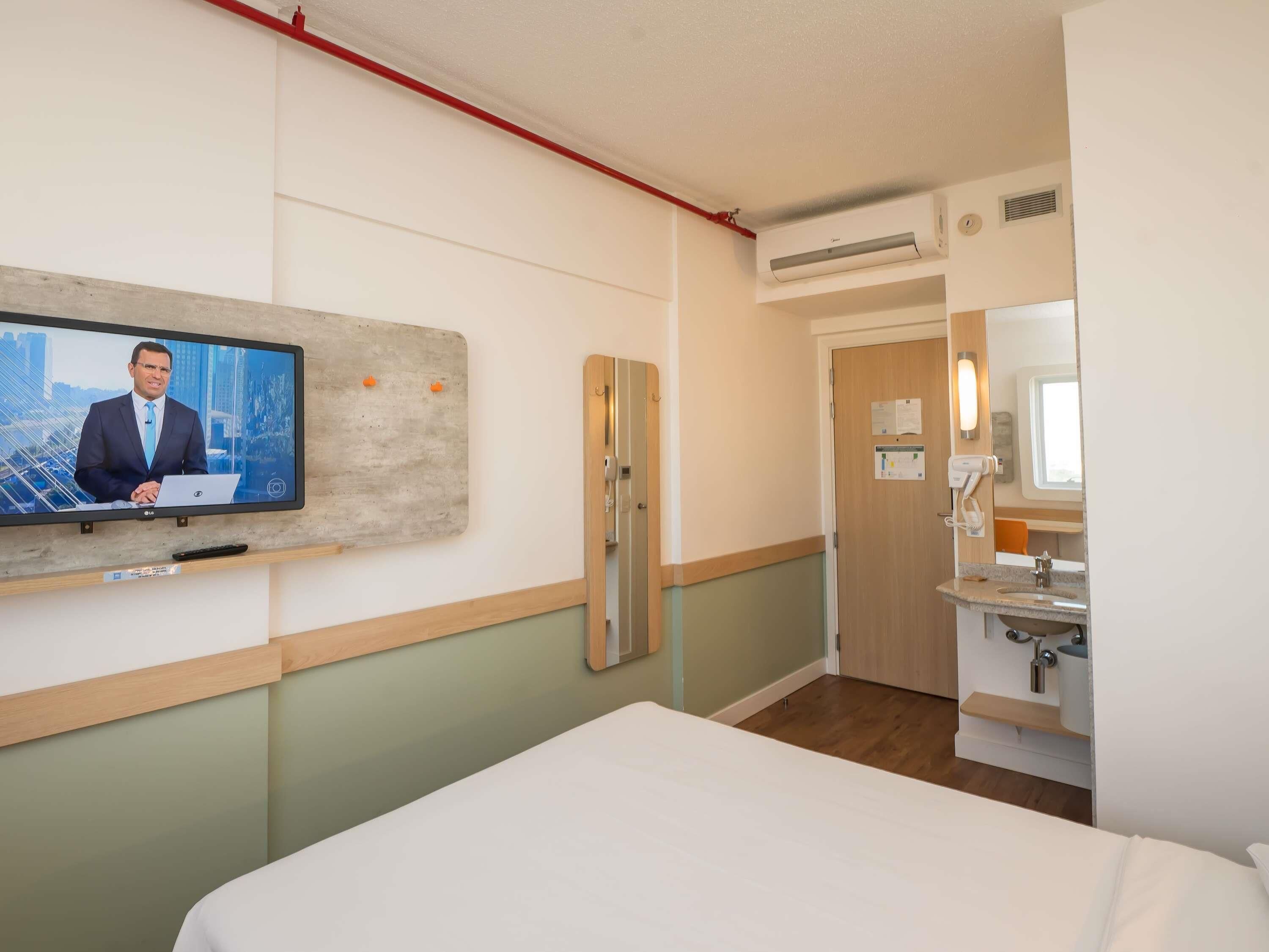 A hotel room with a bed, a TV mounted on the wall, a wooden door, a bathroom with a sink and mirror, and an air conditioning unit.