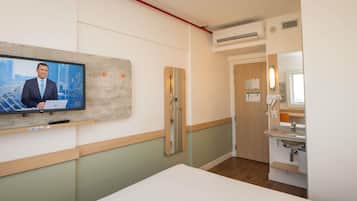 A hotel room with a bed, a TV mounted on the wall, a wooden door, a bathroom with a sink and mirror, and an air conditioning unit.