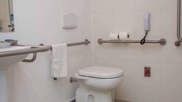 Casa de banho com sanita, barra de apoio, lavatório e suporte para rolo de papel higiénico.