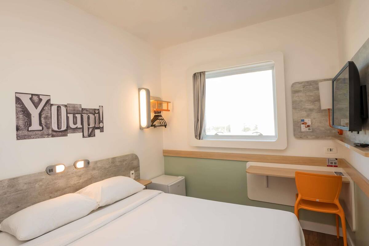 Une chambre d’hôtel équipée d’un lit, d’un bureau, d’une chaise, d’une télévision, d’une fenêtre et d’un élément décoratif mural.