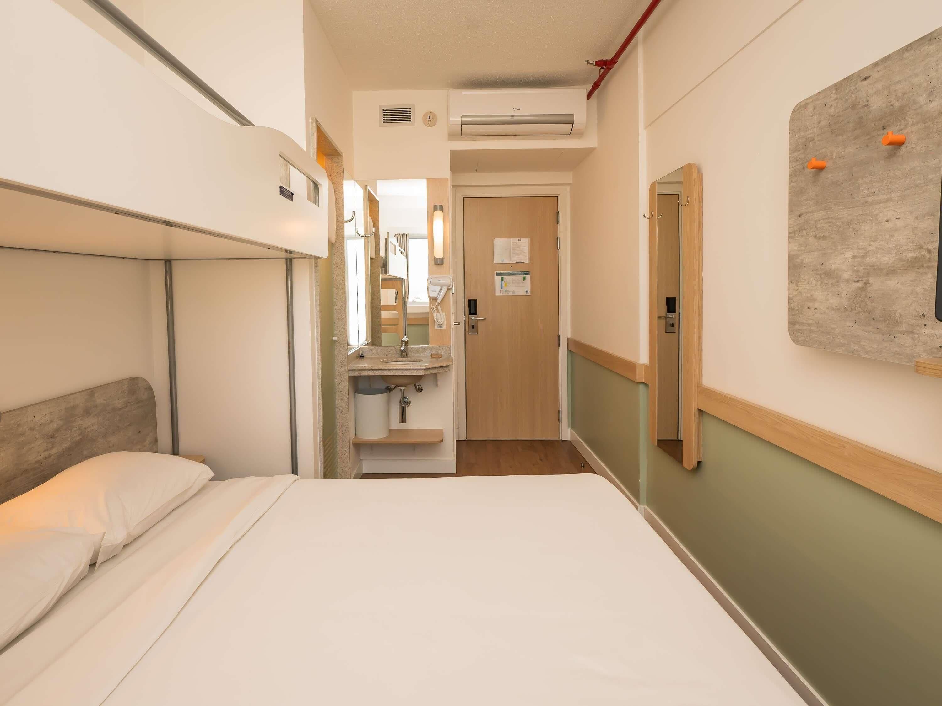 Quarto de hotel com beliche, banheiro com pia, espelho e dispensador de toalhas, e porta que dá para outro quarto.