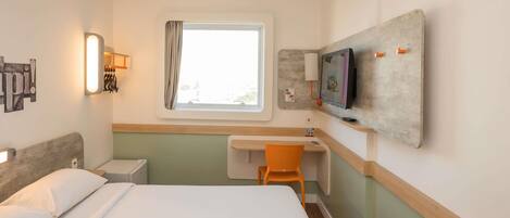 Habitación de hotel con cama, escritorio, silla, televisor montado en la pared y ventana con cortinas.