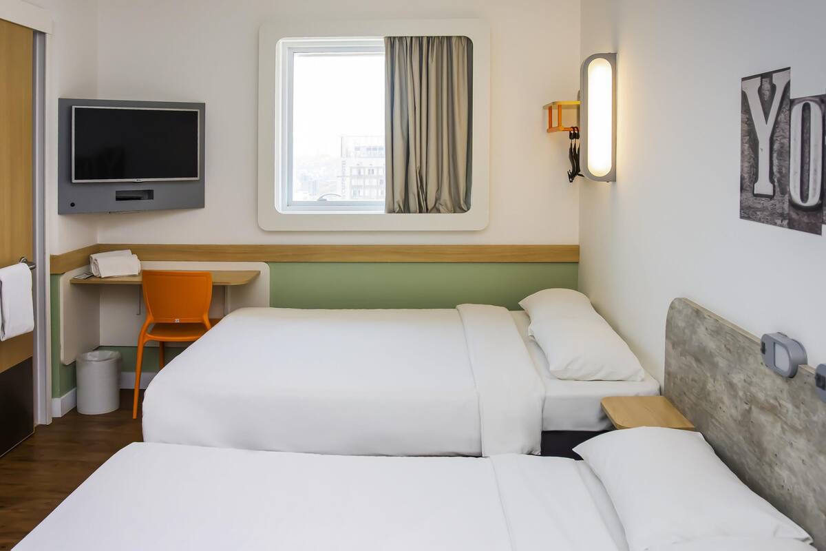 Une chambre d’hôtel avec deux lits, une télévision, un bureau avec une chaise, une fenêtre avec des rideaux et un luminaire fixé au mur.