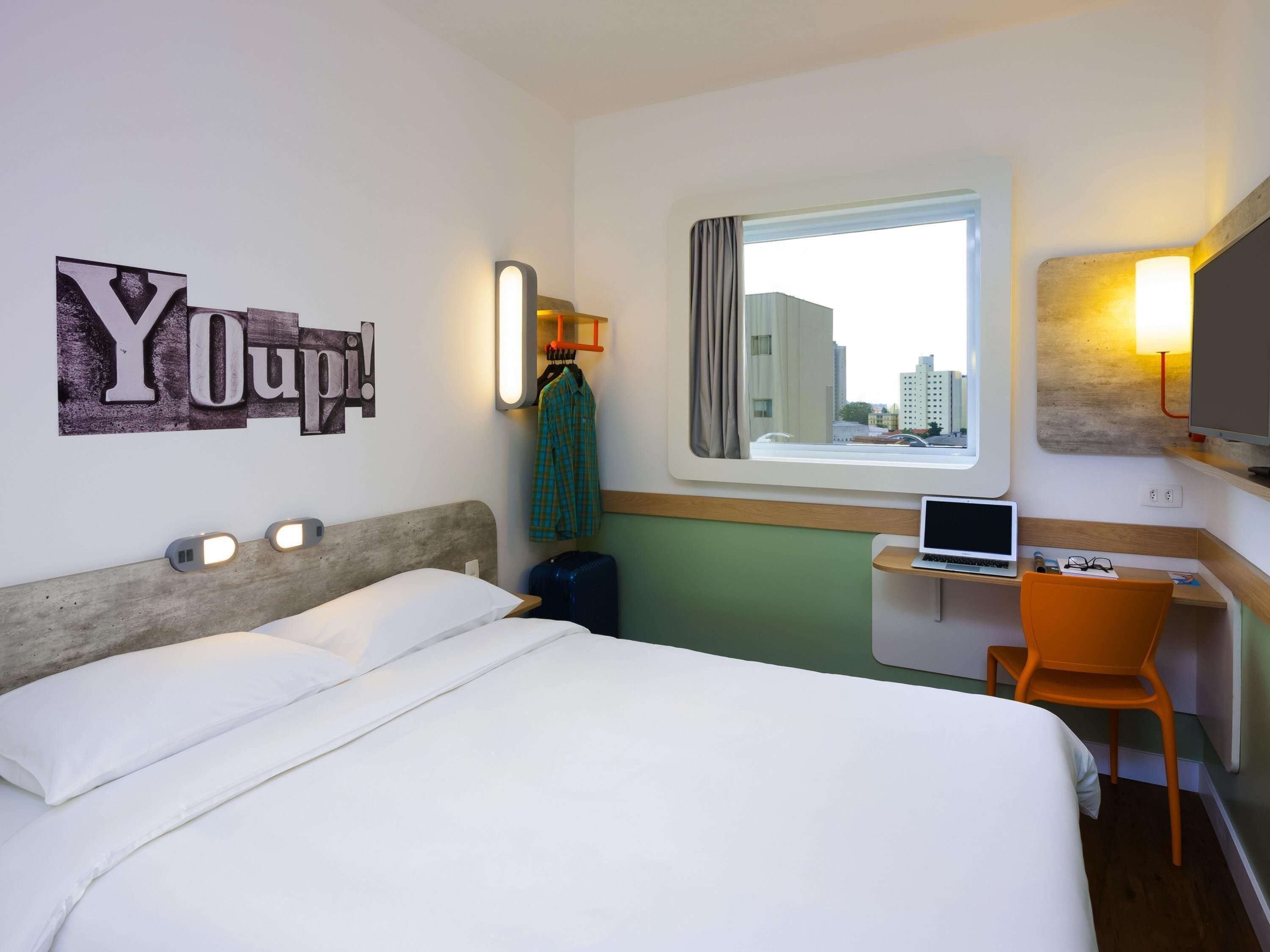Une chambre d’hôtel avec un lit, un bureau avec un ordinateur portable, une chaise, une fenêtre donnant sur des immeubles et un mur orné d’une affiche « YOUP! ».