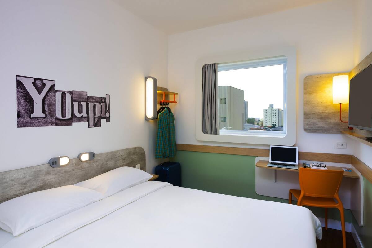 Une chambre d’hôtel avec un lit, un bureau avec un ordinateur portable, une chaise, une fenêtre donnant sur des immeubles et un mur orné d’une affiche « YOUP! ».