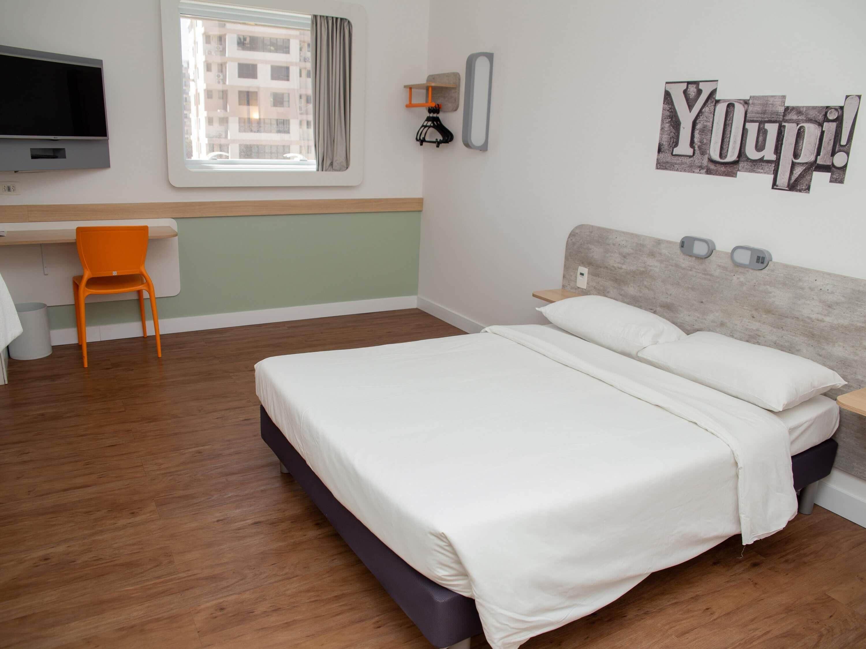 A bedroom with a bed, a desk, a chair, a TV, and a window with curtains.
