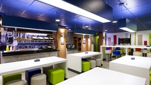 A modern cafe with white tables, colorful stools, and a bar area with food display.