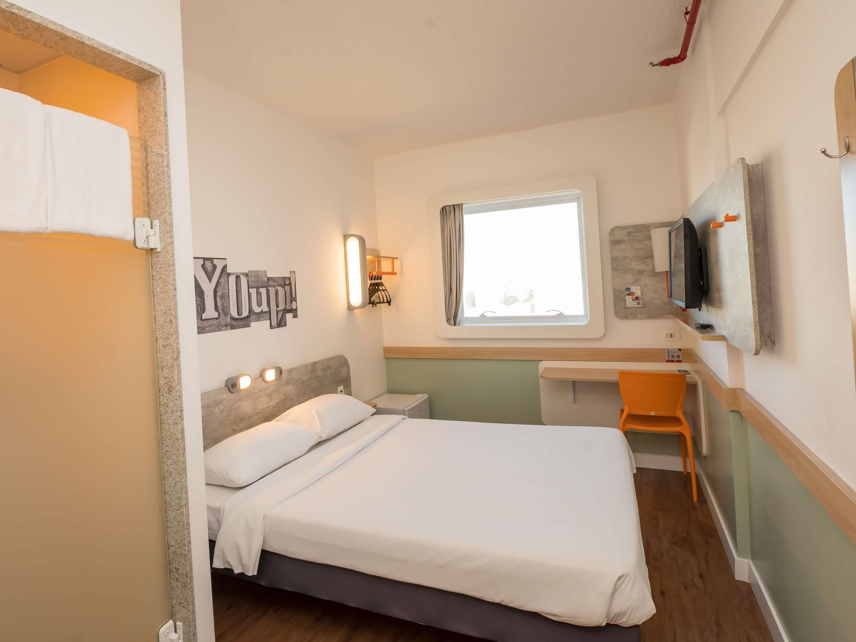 Habitación de hotel con una cama, un escritorio, un televisor fijado a la pared y un ventanal con cortinas.