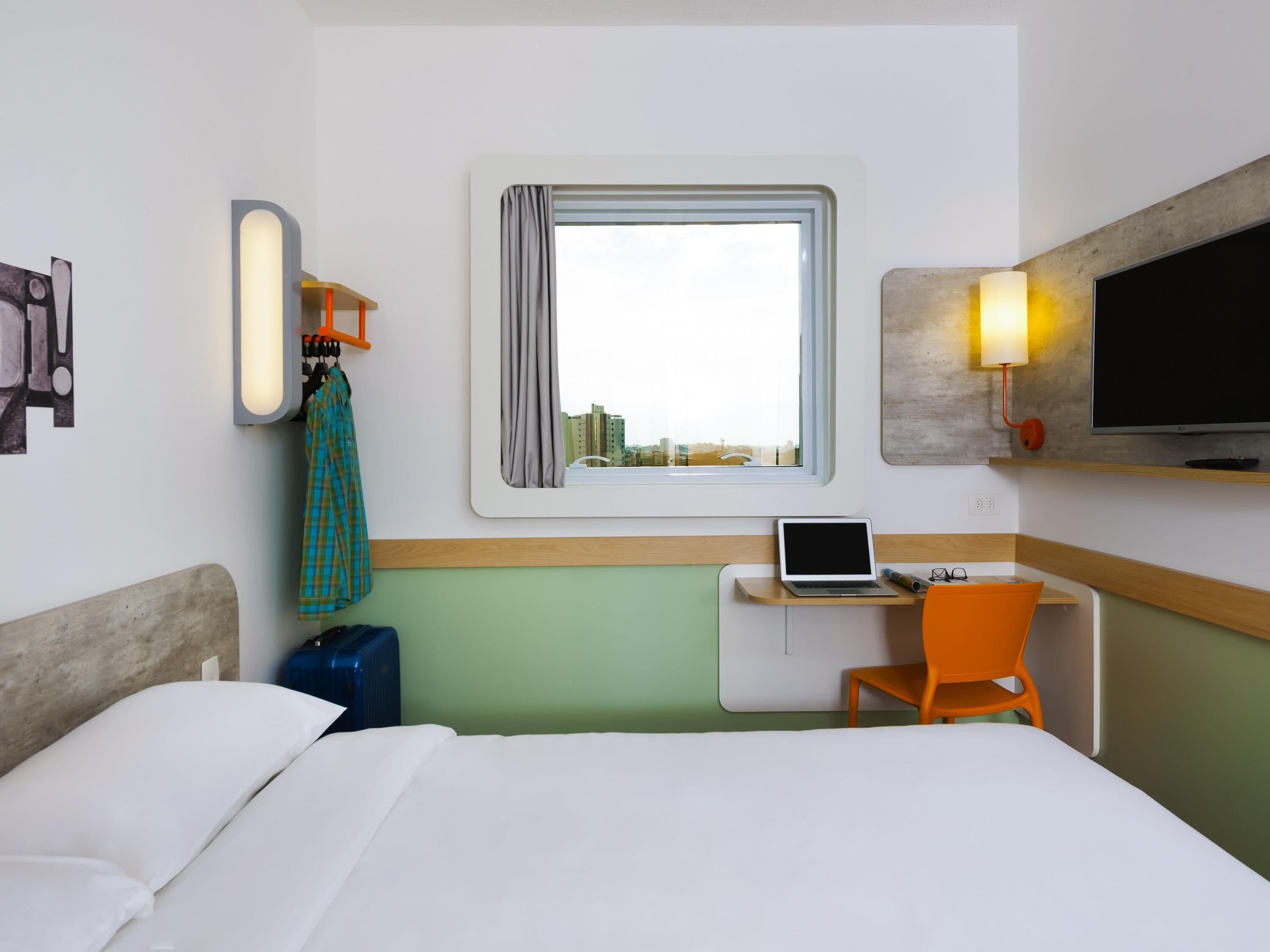 Une chambre d’hôtel avec un lit, un bureau avec un ordinateur portable, une chaise, une valise, une télévision et une fenêtre donnant sur des immeubles.
