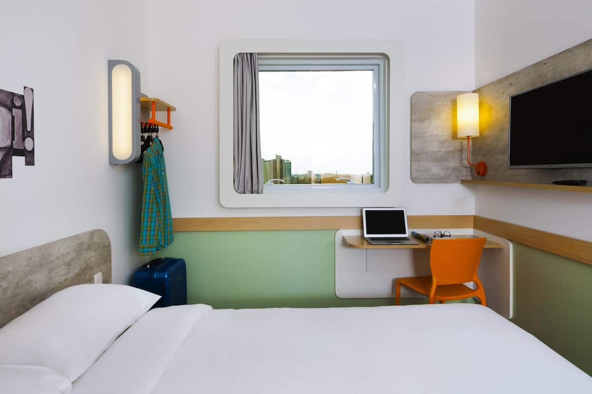 Hotellihuone, jossa on sänky, työpöytä kannettavalla tietokoneella, tuoli, matkalaukku, televisio ja ikkuna, josta on näkymä rakennuksiin.
