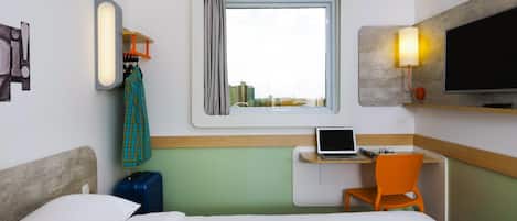 Habitación de hotel con cama, escritorio con computadora portátil, silla, maleta, televisión y ventana con vista a edificios.