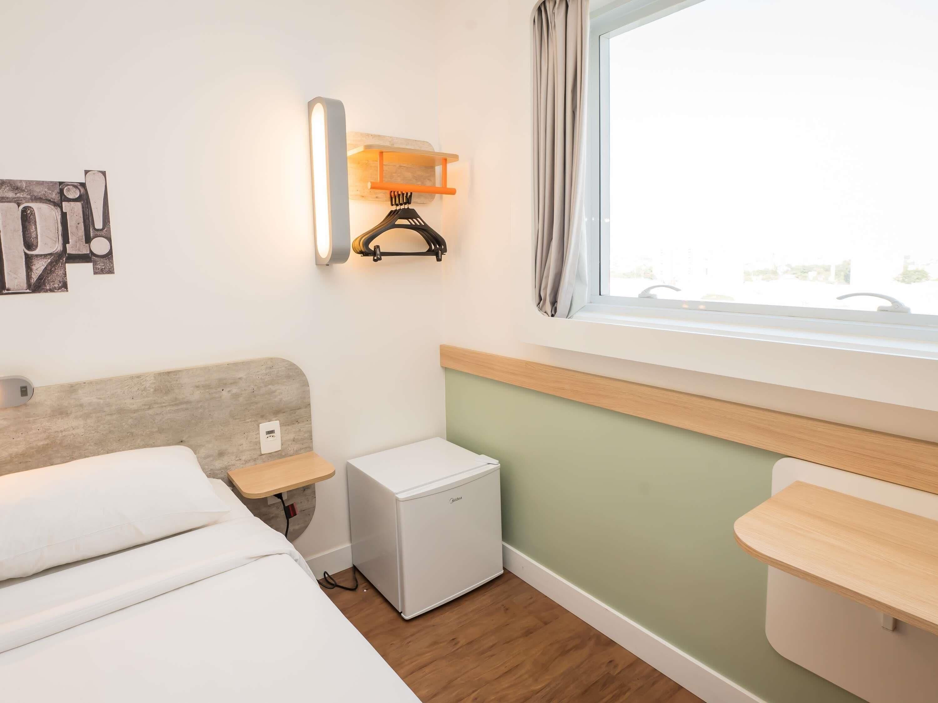 Quarto de hotel com cama, mesinha de cabeceira pequena, mini-frigorífico, luminária na parede, janela com cortinas e porta-toalhas.