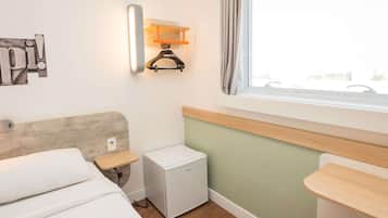 Quarto de hotel com cama, mesinha de cabeceira pequena, mini-frigorífico, luminária na parede, janela com cortinas e porta-toalhas.