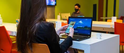Una persona usando un teléfono inteligente en una mesa blanca en una habitación con paredes verdes, una computadora portátil y un televisor al fondo.