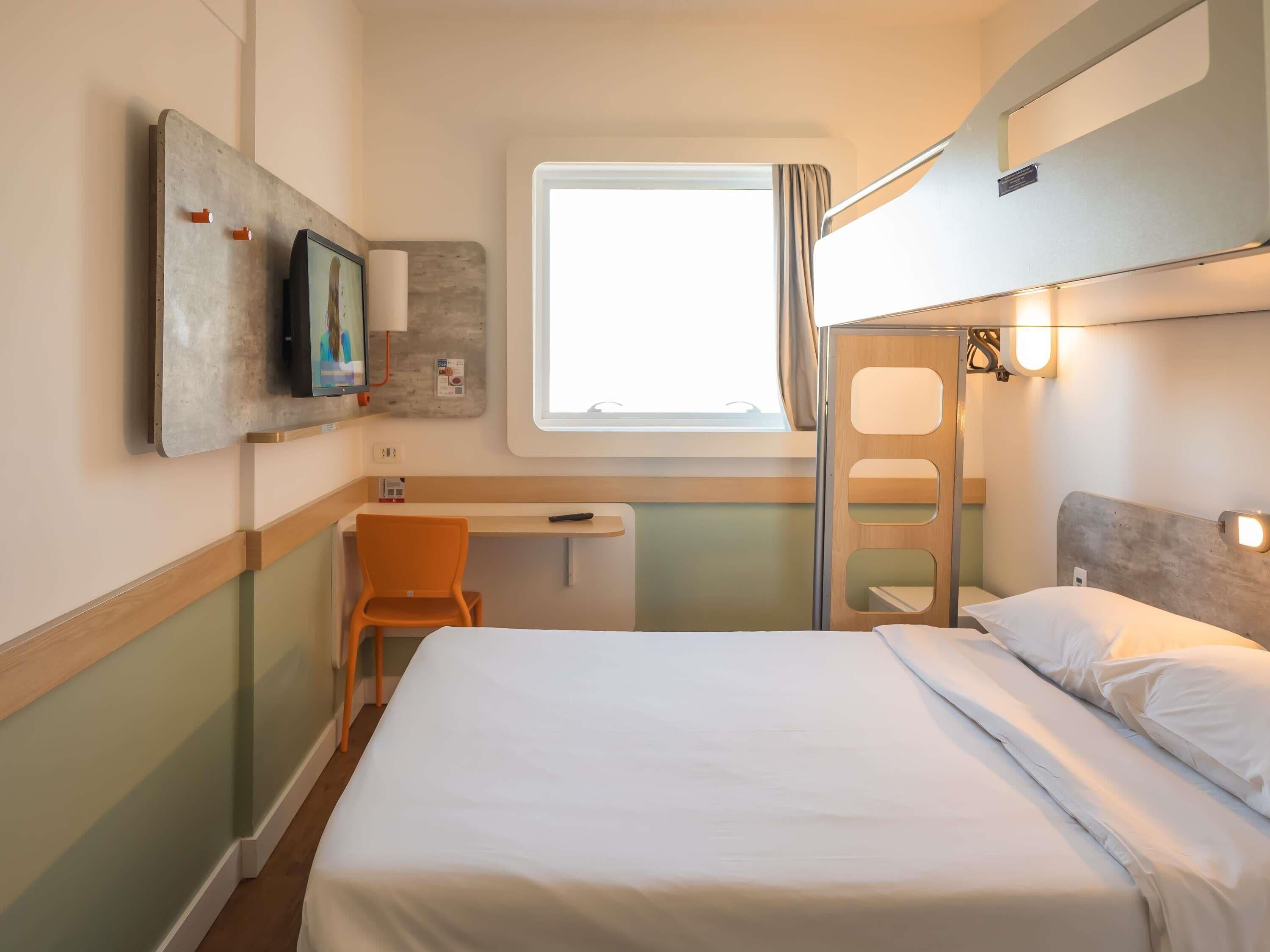 A compact hotel room with a bed, a desk, an orange chair, a TV, and a window.