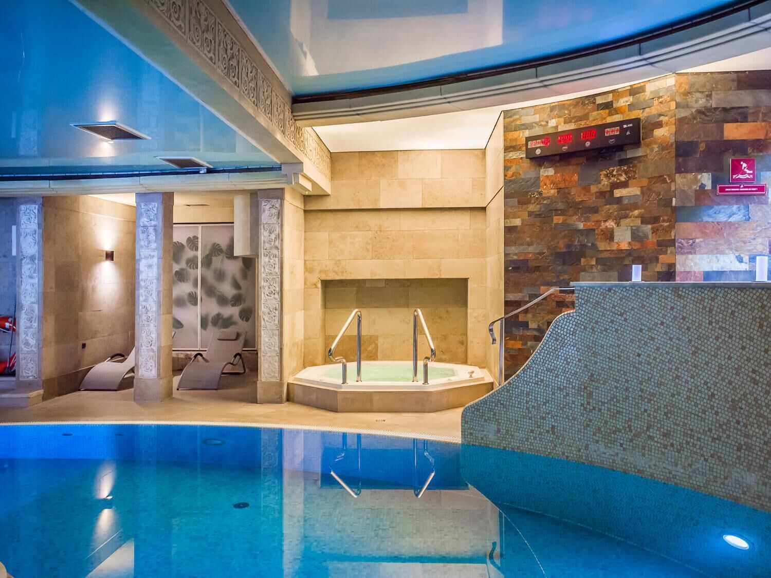 Piscina interior moderna com hidromassagem, parede de pedra e relógio digital.
