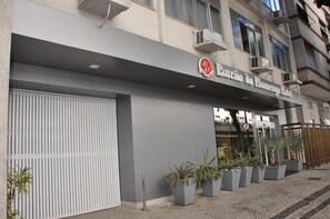 Entrada de hotel con una puerta blanca cerrada, un letrero con logotipo rojo y blanco, y plantas en macetas al frente.
