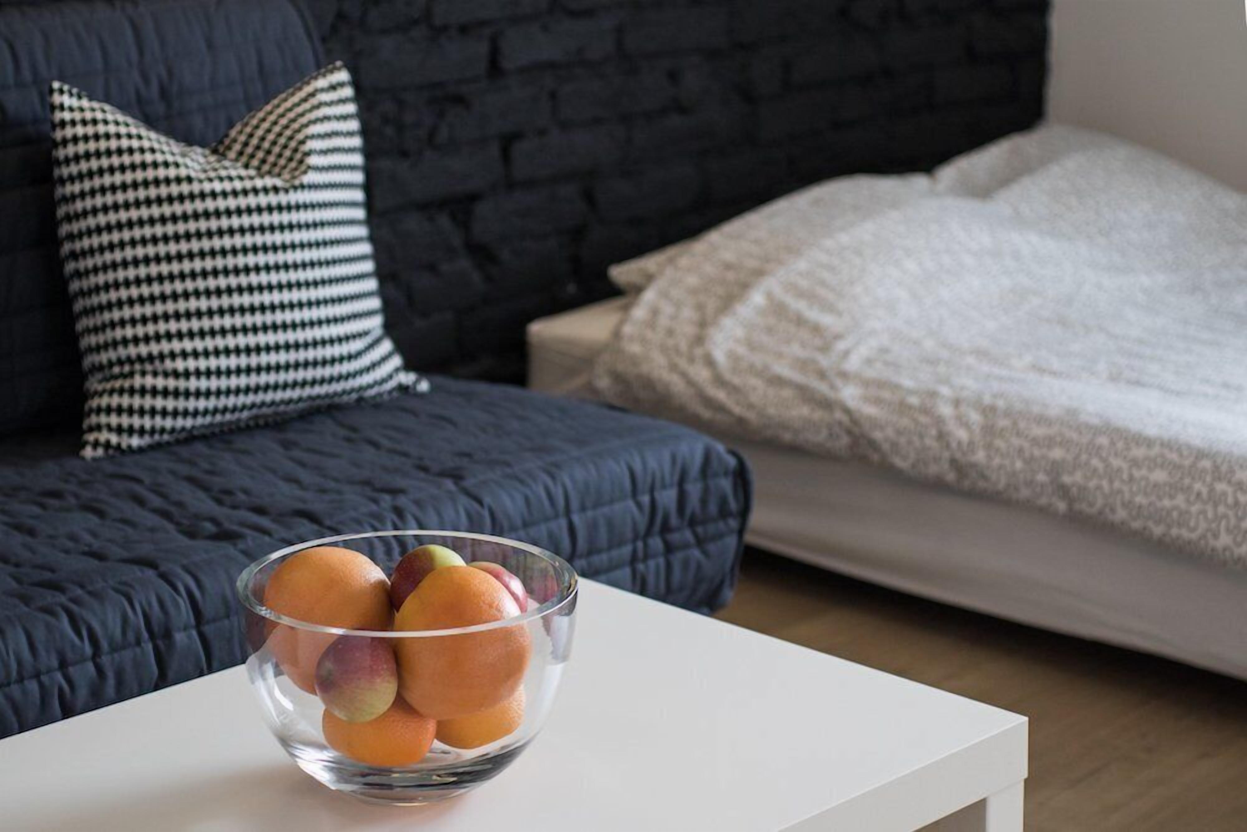 A modern living room with a dark blue sofa, a white coffee table with a fruit bowl, and a contrasting white bed with a textured blanket.