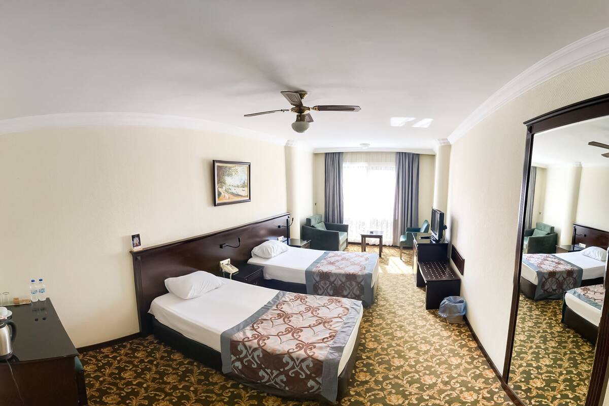 Habitación de hotel con dos camas, ventilador de techo, escritorio, silla, televisor y un espejo grande.