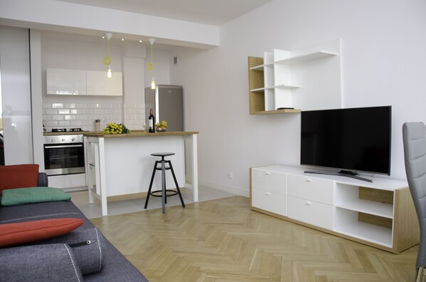 A modern kitchen with white cabinets, a built-in oven, and a refrigerator. There is a dining area with a gray sofa and red cushions. A TV is mounted on a white cabinet, and a bar stool is visible.