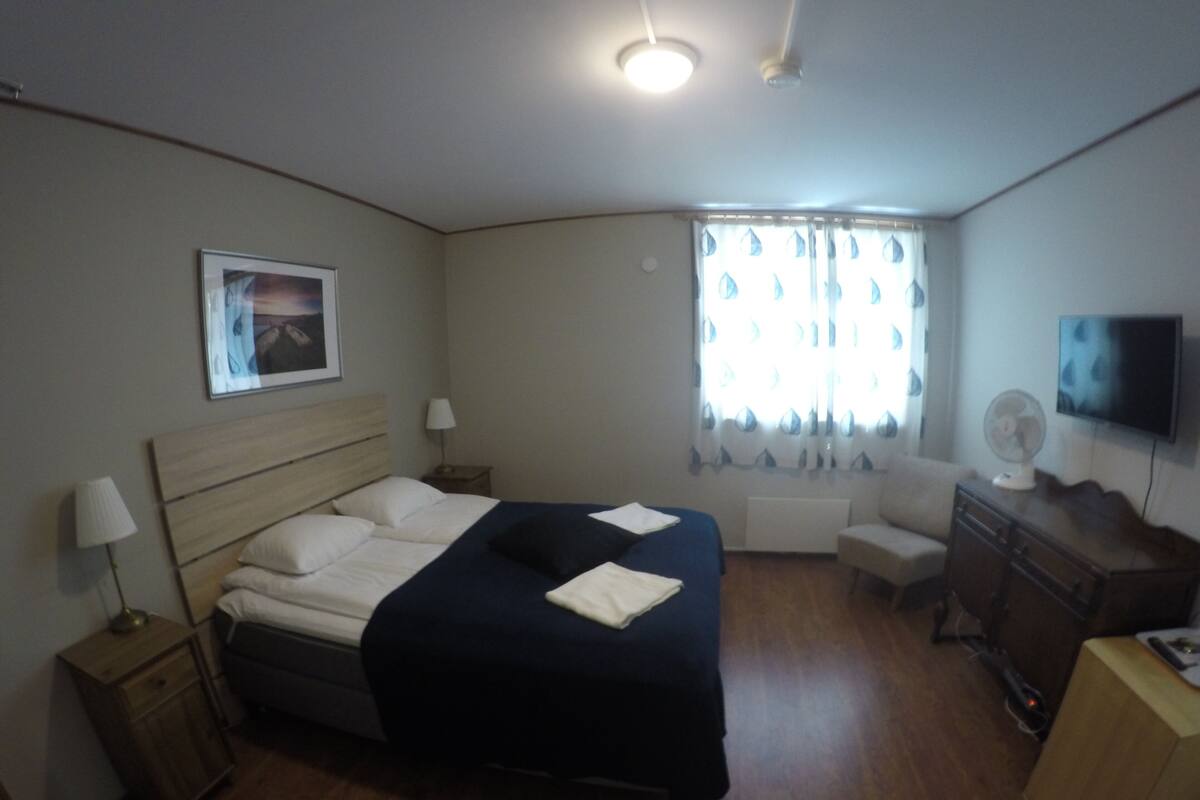 A hotel room with a bed, bedside tables, a desk, a chair, a TV, and a window with curtains.