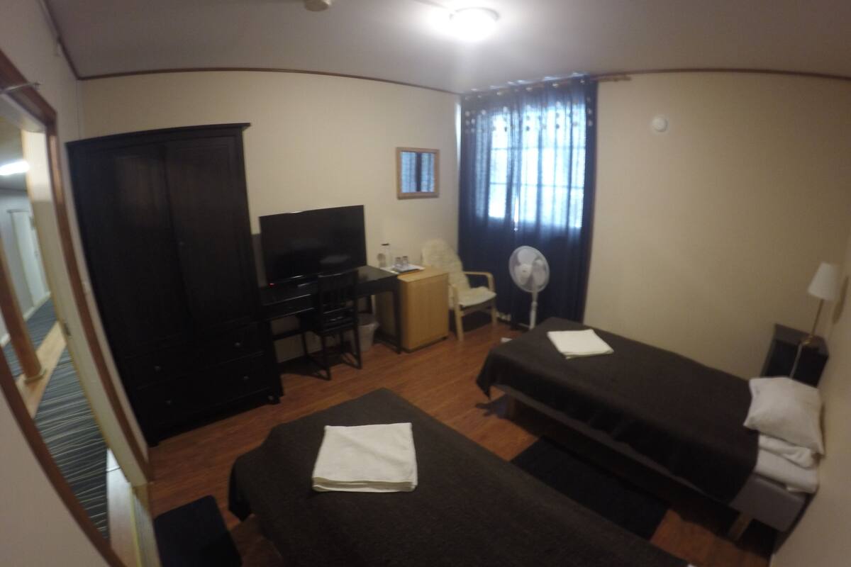 A hotel room with two beds, a desk with a TV, a chair, a fan, and a window with curtains.