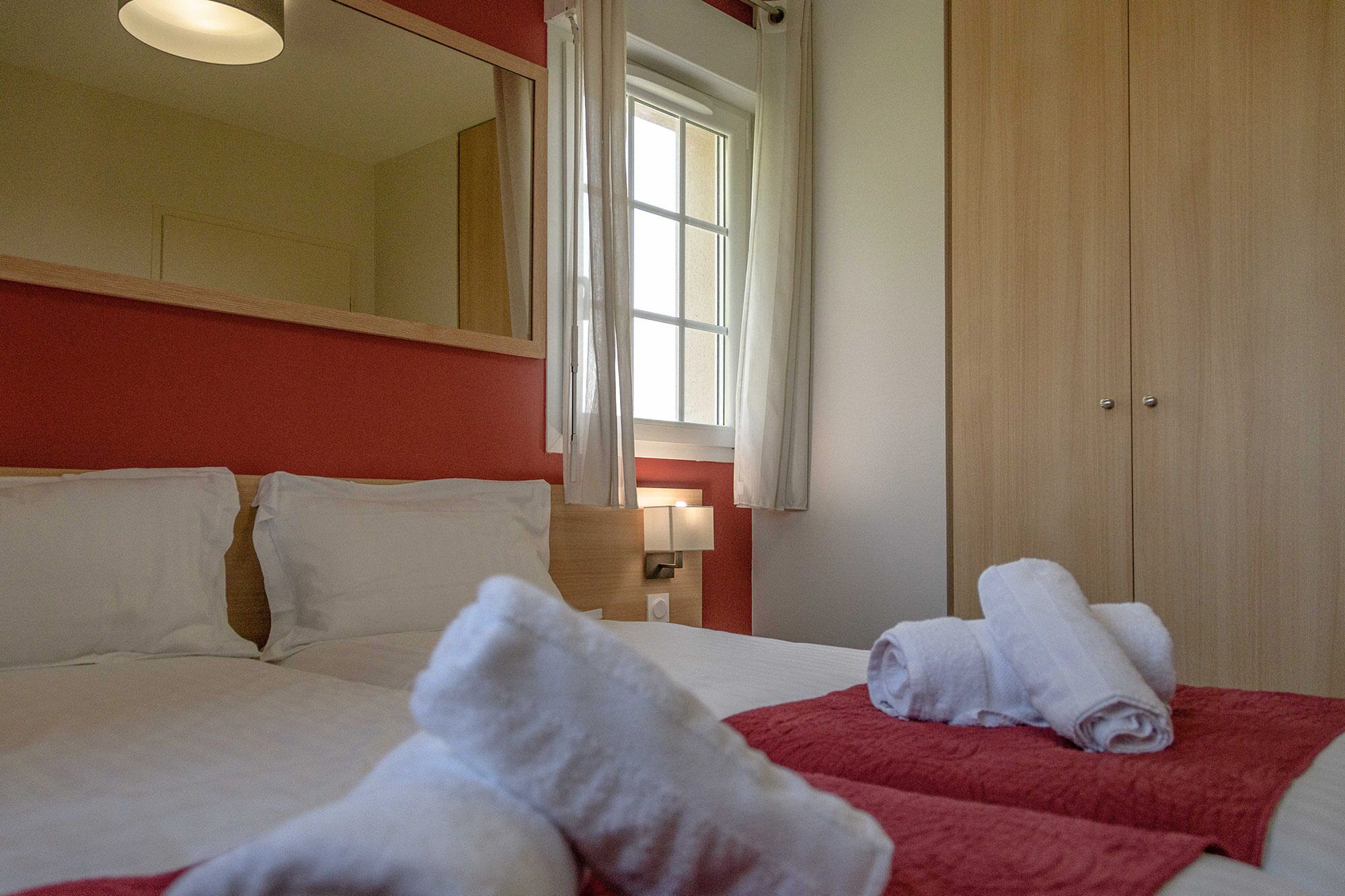 A hotel room with a bed, a red wall, a wooden wardrobe, and a window with curtains.